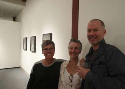 Three adults smiling, standing in an art gallery with framed pictures in the background. they appear cheerful and are casually dressed.