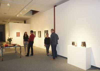 People viewing and discussing artwork at an art gallery exhibition. the space features white walls and is well-lit, with paintings and sculptures displayed.