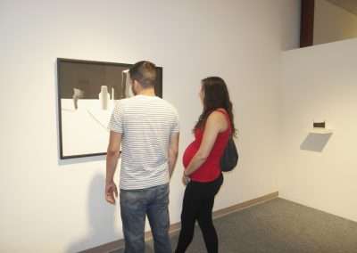 A man and a woman viewing an abstract artwork in a gallery setting. the woman is wearing a red top and has long hair, while the man is in a striped shirt.