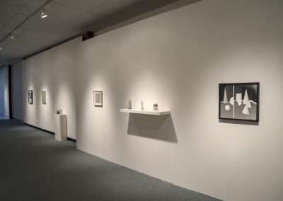 Interior of a modern art gallery with white walls displaying various framed artworks and sculptures on pedestals, illuminated by ceiling lights.