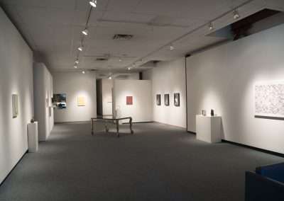 Interior of an art gallery with white walls displaying various paintings and artworks, a seating area with blue chairs, and track lighting overhead.