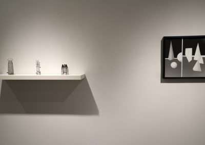 A minimalist art installation featuring a white shelf with assorted abstract sculptures placed against a grey wall, alongside a black framed artwork with geometric shapes.