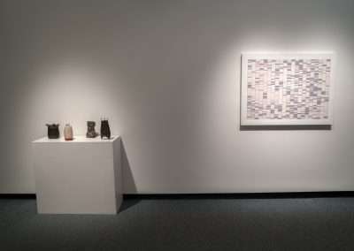 Art gallery interior with three ceramic vases on a white pedestal and a large abstract mosaic art piece displayed on a gray wall, illuminated by soft overhead lighting.