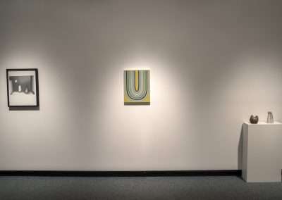 An art gallery wall with three displayed pieces: a framed photograph on the left, a colorful abstract painting in the middle, and a sculpture of a cat on a pedestal on the right.
