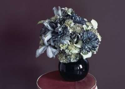 A lush bouquet of dark blue and white flowers in a glossy black vase, placed on a red stool against a deep purple background.
