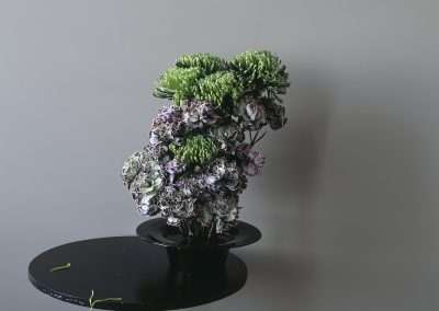 A bouquet of green and purple flowers arranged in a vase on a small black table against a gray background.