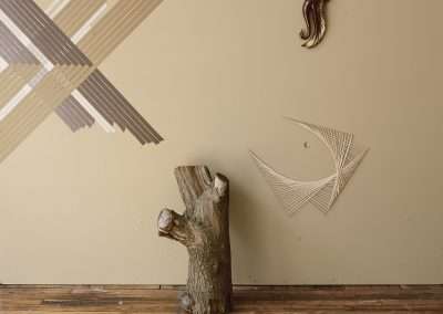 A large piece of wood resembling a tree trunk stands on a wooden floor against a beige wall adorned with abstract geometric tape art and a small nautical-themed decoration.