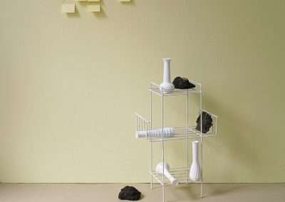 A minimalist setup featuring a white metal shelf with various white vases and dishes, accompanied by black rocks, against a light green wall with yellow sticky notes.