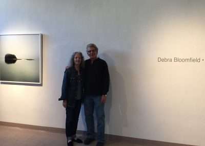A man and a woman standing together in a gallery, smiling next to a wall bearing the name "Debra Bloomfield" and an artwork featuring a serene aquatic scene.