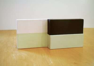 Two minimalist rectangular blocks painted in soft pastel colors, white and pale green with a dark section, resting on a wooden surface.