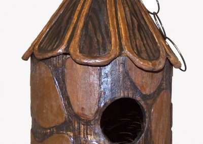 A rustic wooden birdhouse with a conical roof, featuring a circular entrance and embellished with a pattern of lighter wooden patches.