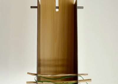 A minimalist wooden sculpture consisting of a tall, vertical, split wooden panel with multiple thin green rods inserted horizontally at the base, creating a layered effect.
