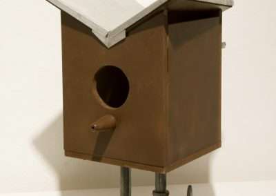 A simple brown wooden birdhouse with a sloping metal roof, circular entrance, and mounted on a metal clamp.