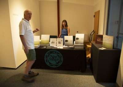 An older man wearing a white shirt and shorts walks past a book display table manned by a young woman in a blue dress. The table features several books and a sign that reads "New Mexico.