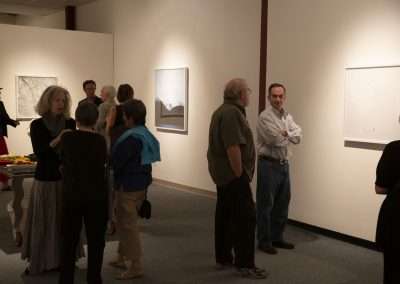 People interacting at an art gallery with various framed artworks on the walls and visitors standing or conversing in groups. The lighting is soft and the mood appears contemplative.