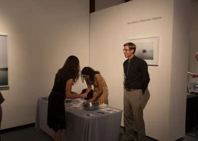 Three people interact at a gallery event beside an information booth labeled "New Mexico Wilderness Alliance." One man is standing while two women are engaged with materials on the table.