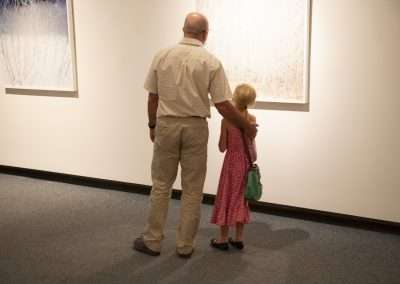 An older man and a young girl, possibly grandfather and granddaughter, view framed abstract artwork in a gallery, standing side by side with a warm embrace.