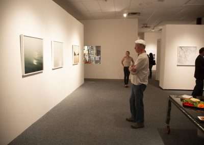 An art gallery with white walls displaying various framed artworks. Two men, one in a white cap, stand talking near a food table. A woman in the background observes the art.
