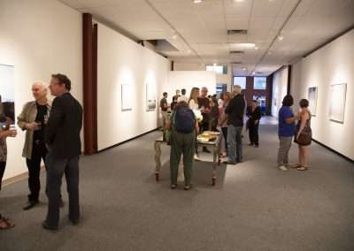 People socializing at an art gallery opening, viewing artworks on the walls and conversing with drinks in hand. A food cart is also visible in the scene.