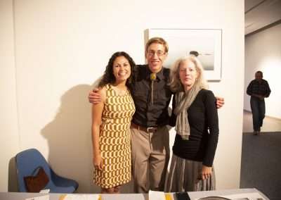 Three people smiling, two women and one man, standing together at an indoor event, brightly lit, with art on the walls and a blue chair to the left.
