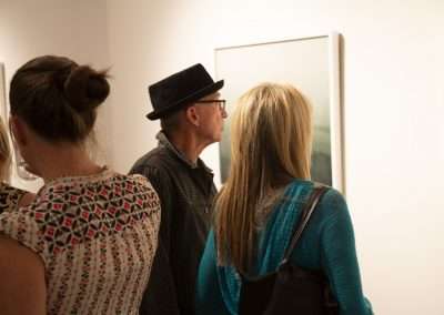 People viewing artwork in a gallery; the image captures a man in a hat and two women examining framed pieces on the wall.