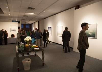 A gallery opening with people viewing large framed photographs on white walls, and a man in a dark blazer walking thoughtfully past a refreshment table.