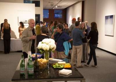A lively art gallery reception with groups of people conversing. A table in the foreground displays an arrangement of flowers and refreshments. Artwork is visible on the walls in the background.