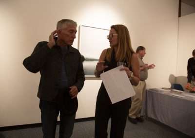 Two people having a conversation in an art gallery, with one person holding papers and the other adjusting their hearing aid. More visitors are visible in the background.