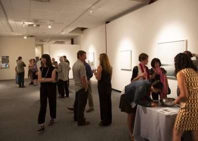People interact and view art at an exhibition in a gallery with white walls and spotlights. Visitors are standing, talking, and examining artworks, while one person signs a guest book at a table.