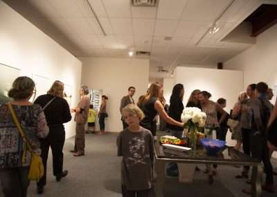 People mingling at an art gallery exhibition featuring large paintings, with a central table displaying flowers and refreshments.