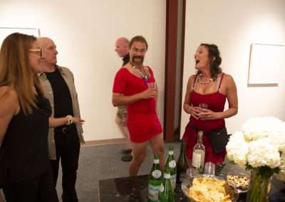 A lively group of adults chat at an indoor party, with a woman in a red dress laughing joyously. A table with snacks and drinks is in the foreground.