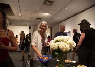 People gathered at an art gallery event, with a lady in the foreground reaching for snacks, others conversing in the background, surrounded by artwork.