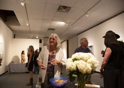 An art gallery scene with people mingling. A woman with gray hair stands in the foreground smiling, holding a wine glass, while other guests chat in the background.