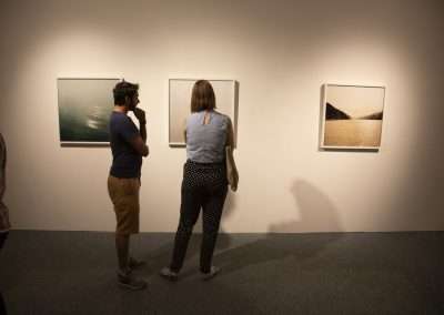 Two people observing photographs in an art gallery, standing in front of three framed pictures of landscapes hung on a well-lit wall.