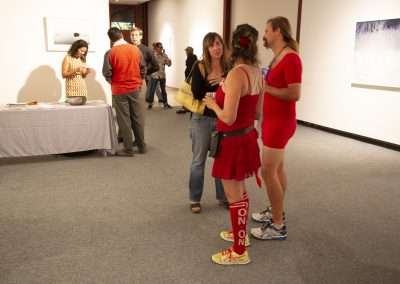 People conversing at an art gallery event, with one woman dressed in sporty red attire, including knee-high socks, standing out in the casual setting.