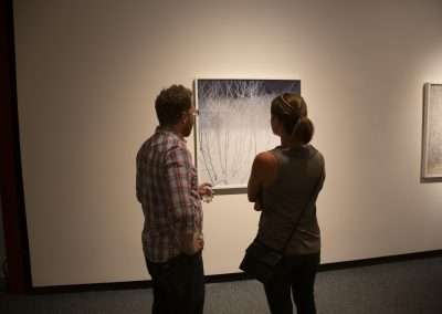 Two people observing a painting depicting bare trees in a gallery, standing close together in discussion, with other artworks visible in the background.