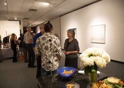 A woman and a man converse in an art gallery filled with guests, artwork on the walls, and a table adorned with flowers and snacks in the foreground.