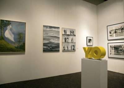 Interior of an art gallery featuring various framed paintings on white walls, and a yellow sculpted "yo" in the foreground.