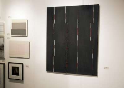 A modern art gallery displaying a large black canvas with vertical red lines, surrounded by smaller framed artworks on white walls.