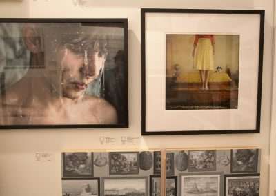 Photographs displayed on a gallery wall; one features a close-up of a crying boy, another showcases a woman in a yellow dress with a dog on a bed. other images visible below.