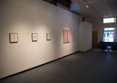An art gallery interior showing a dimly lit room with four framed artworks on a white wall and two blue chairs positioned near the corner.