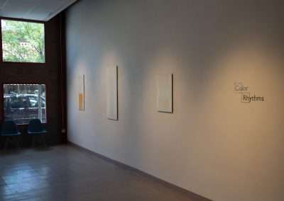 An art gallery interior featuring a wall with four framed artworks and a title "Color Rhythms" on the wall. To the left, two blue chairs face a glass window reflecting trees.