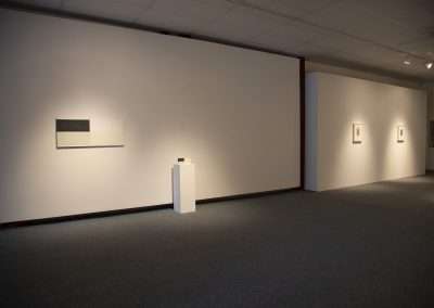 An art gallery interior with dim lighting showing three framed artworks on white walls and a small sculpture on a pedestal.