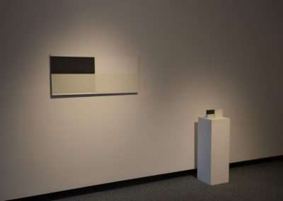 A minimalist art display featuring a simple, two-toned rectangular painting on a gray wall above a short white pedestal with a small black object displayed. Dimly lit ambiance focuses attention on the artwork.