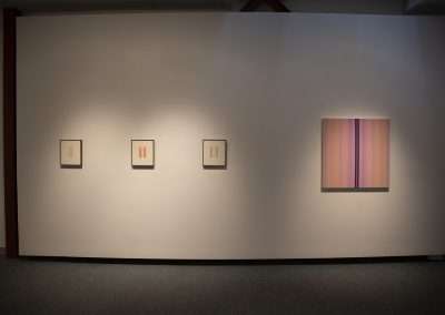 An art gallery wall with four framed pieces displayed under soft lighting, three featuring minimalist designs to the left and one with vertical color stripes to the right.