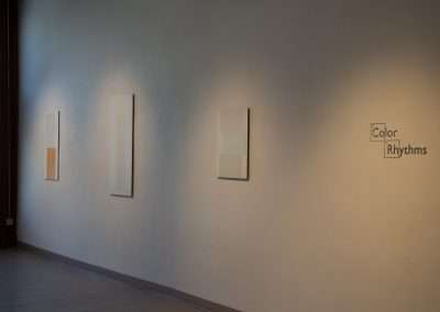Three minimalist paintings, featuring soft color blocks, are displayed on a beige wall of a gallery under soft lighting; labeled "Color Rhythms".