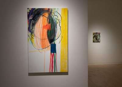 An abstract painting displayed in an art gallery, featuring bold black lines and vibrant colors including yellow, red, and blue on a white background. A smaller painting is visible in the background to the right.