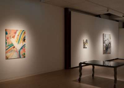 An art gallery room with dim lighting showcasing three abstract paintings on white walls, and a reflective table in the foreground.