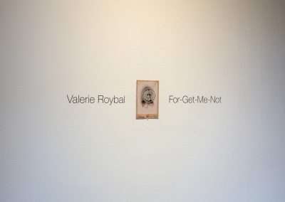 A small framed artwork titled "For-Get-Me-Not" by Valerie Roybal is displayed centered on a white gallery wall, with the artist's name and artwork title printed above and to the left.
