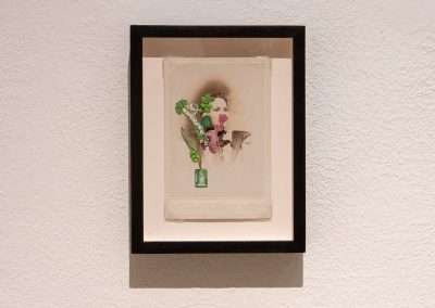 Artwork in a black frame mounted on a white wall, featuring an abstract collage with a face, floral elements, and vibrant colors on an aged paper background.